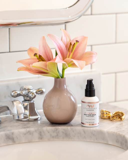 Pictured: a TOUCH N GO™ Daily Hair Serum bottle by a bathroom sink with flowers and earrings beside it.