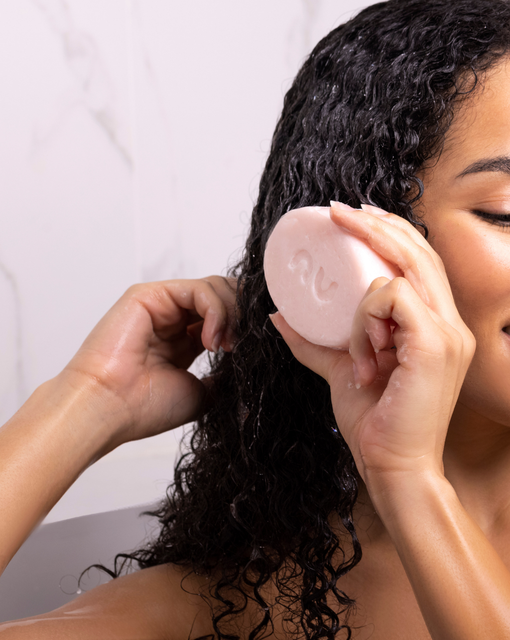 Femme in shower holding the WASH N GO™ Butter Bar Shampoo in her hand. She is holding the product to her hair to show how to apply the bar to wet hair by gliding it along the hair shaft.
